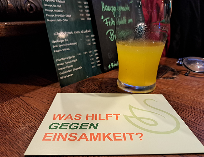 Ein Foto zeigt ein Glas mit Limonade auf einem Tisch in einem Irish Pub. Davor liegt eine Postkarte mit dem Text: "Was hilft gegen Einsamkeit?"